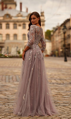 A woman wearing the La Femme 31717 Long Sleeve Mauve Formal Gown with floral embroidery stands on a cobblestone street, facing away and glancing back over her shoulder.