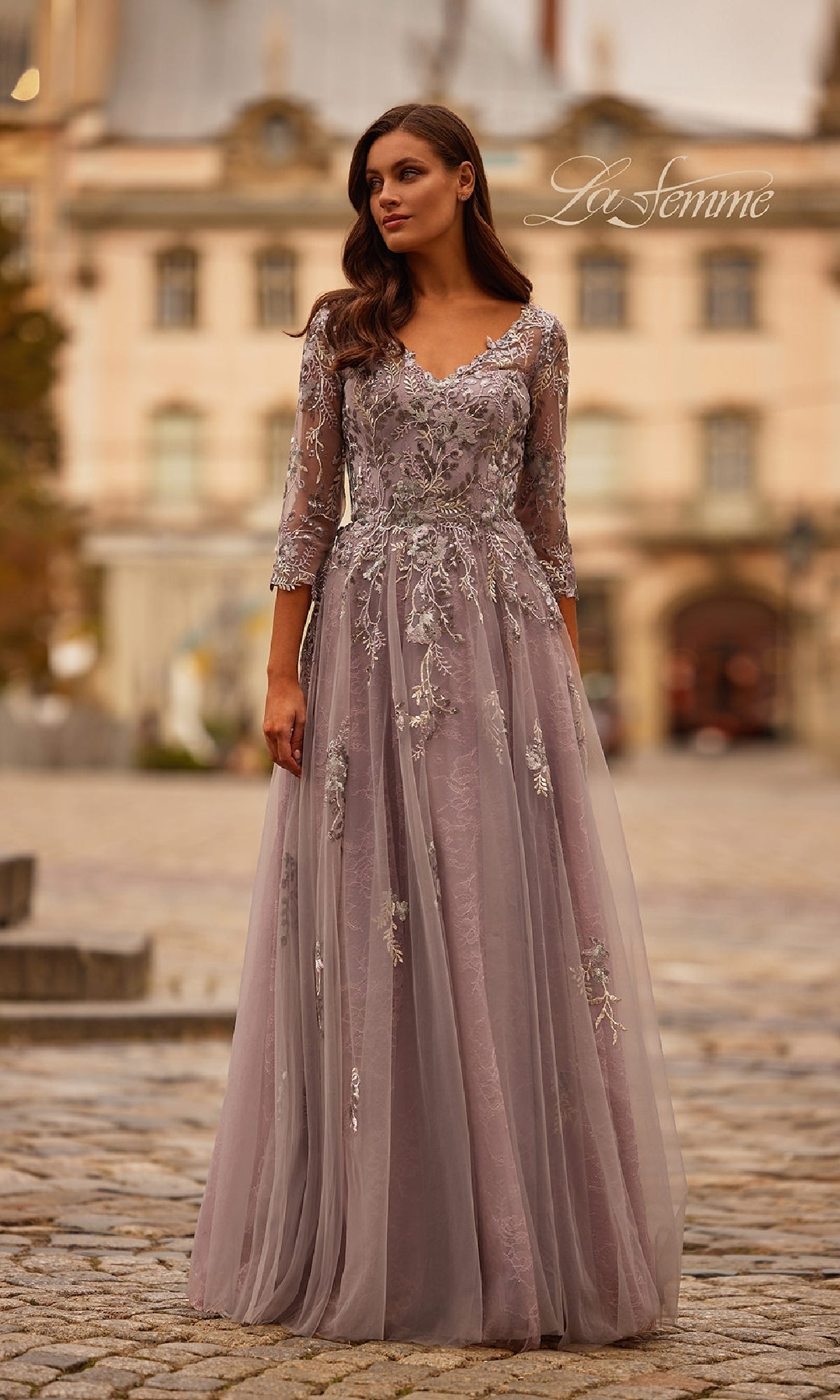 A woman in the Long Sleeve Mauve Formal Gown: La Femme 31717, featuring sheer sleeves and delicate embroidery, stands outdoors on a cobblestone street with historic buildings behind her.