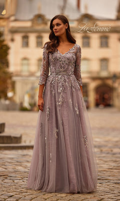 A woman in the Long Sleeve Mauve Formal Gown: La Femme 31717, featuring sheer sleeves and delicate embroidery, stands outdoors on a cobblestone street with historic buildings behind her.