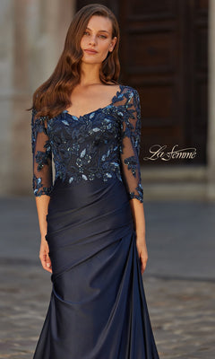 A woman stands outdoors on a cobblestone path wearing the La Femme 31802 long formal dress—a navy blue satin gown with a sheer, sequined beaded lace bodice and three-quarter sleeves.