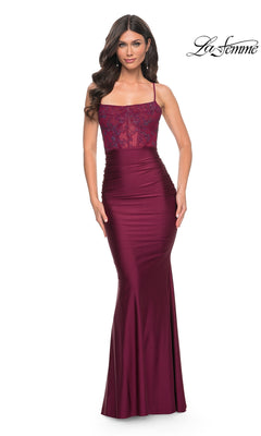 A woman wearing the La Femme Sheer-Lace-Corset Long Prom Dress 31857 in Dark Berry, featuring a fitted, floor-length silhouette with a sheer, embroidered bodice and thin straps, stands against a white background.