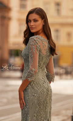 A woman in the La Femme 31909 long formal dress, featuring light green lace, three-quarter sleeves, and sheer details, stands outdoors looking over her shoulder against an elegant city backdrop.