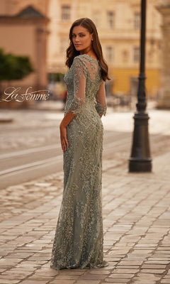 A woman stands on a cobblestone street in the La Femme 31909, a sage green lace long formal dress with sheer three-quarter sleeves and floral embroidery, looking back over her shoulder.