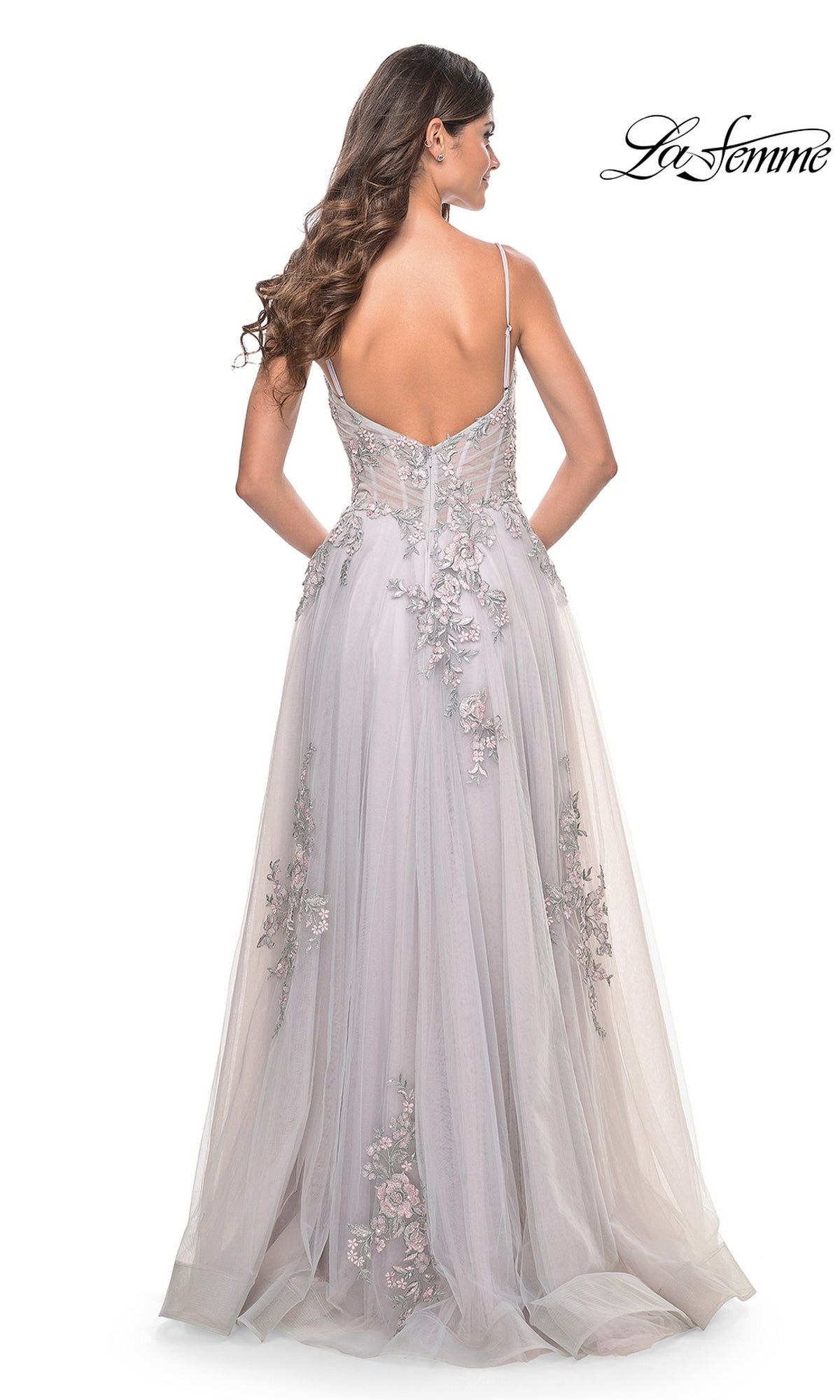 A woman with long wavy hair models the La Femme Embroidered-Lace Long Prom Dress 31939, a light gray, floor-length gown with thin straps and an open back, standing against a white background.
