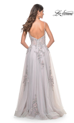 A woman with long wavy hair models the La Femme Embroidered-Lace Long Prom Dress 31939, a light gray, floor-length gown with thin straps and an open back, standing against a white background.