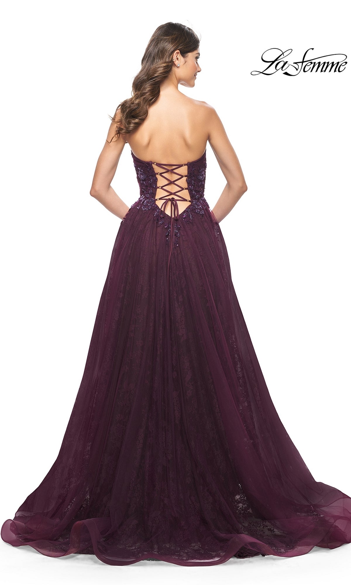A woman wearing the La Femme Strapless Long Prom Ball Gown 31954—a floor-length deep purple dress with a lace-up corset back and sheer skirt—stands facing away.