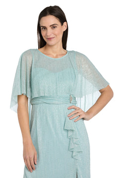 A woman in the R&M Richards 3195P Caped Knee-Length Party Dress, featuring light blue textured fabric, loose short sleeves, and a decorative waist detail, stands with one hand on her hip.