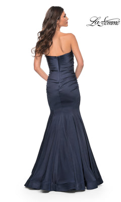 A woman with long brown hair wears the La Femme Strapless Long Mermaid Prom Dress 31980, a navy blue, floor-length gown with subtle ruching and a fitted bodice, shown from the back.