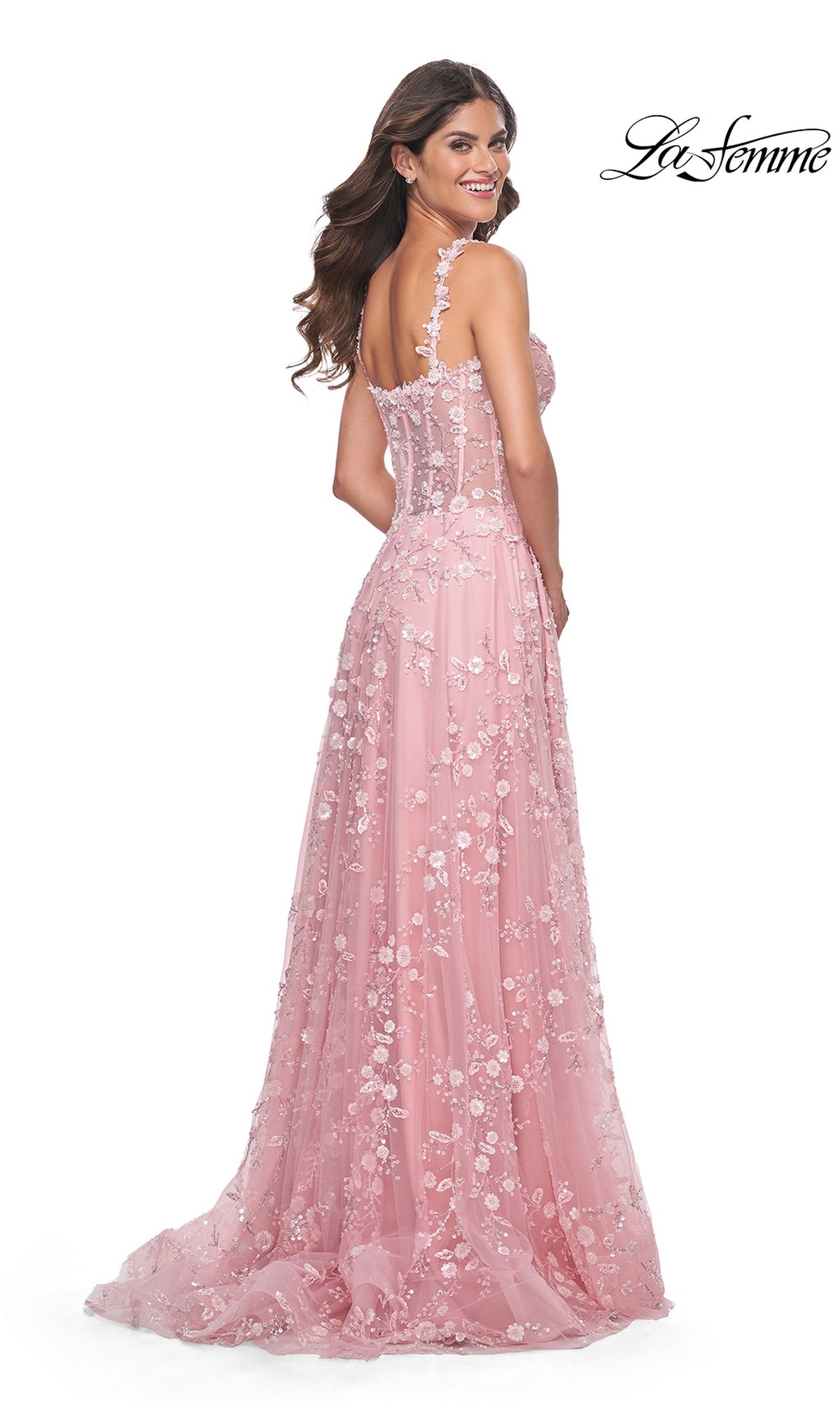 A woman faces backward and smiles in the La Femme Floral-Embellished Long Prom Dress 31996, a sheer pink gown with floral accents. 