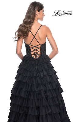 A woman wears the La Femme Long Ruffled Prom Ball Gown 32002, a black A-line designer dress with thin straps, a lace-up back, fitted bodice, and voluminous ruffled skirt. The La Femme logo appears in the top right corner.