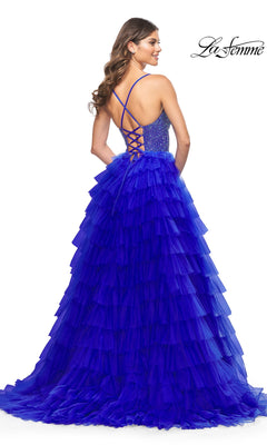 Woman wearing the La Femme Long Ruffled Prom Ball Gown 32002 in royal blue, featuring a lace-up open back and dramatic ruffles, poses with her back to the camera. "La Femme" logo is displayed in the top right corner.