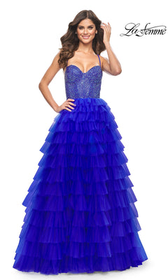 A woman smiles in the La Femme Long Ruffled Prom Ball Gown 32002—a sleeveless, floor-length blue dress with a fitted beaded bodice and tiered ruffled skirt. The logo in the top right highlights this elegant gown.
