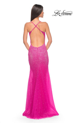 A woman facing away wears the La Femme Sheer-Waist Long Beaded Prom Dress 32007, a bright pink, floor-length, backless gown featuring crisscross straps and sparkling rhinestone embellishments.