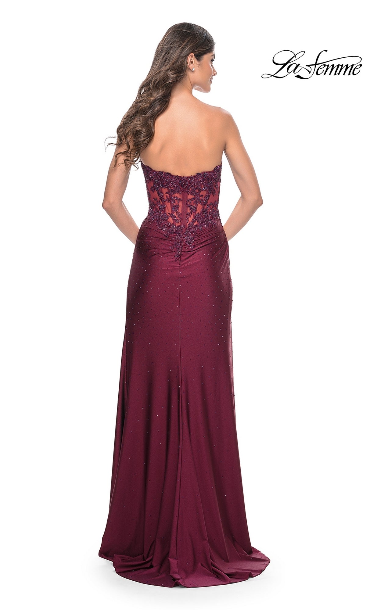 A woman with long wavy hair wears the La Femme Sheer-Waist Strapless Long Prom Dress 32011 in burgundy, with a sheer lace bodice and fitted silhouette. She faces away from the camera, and the 