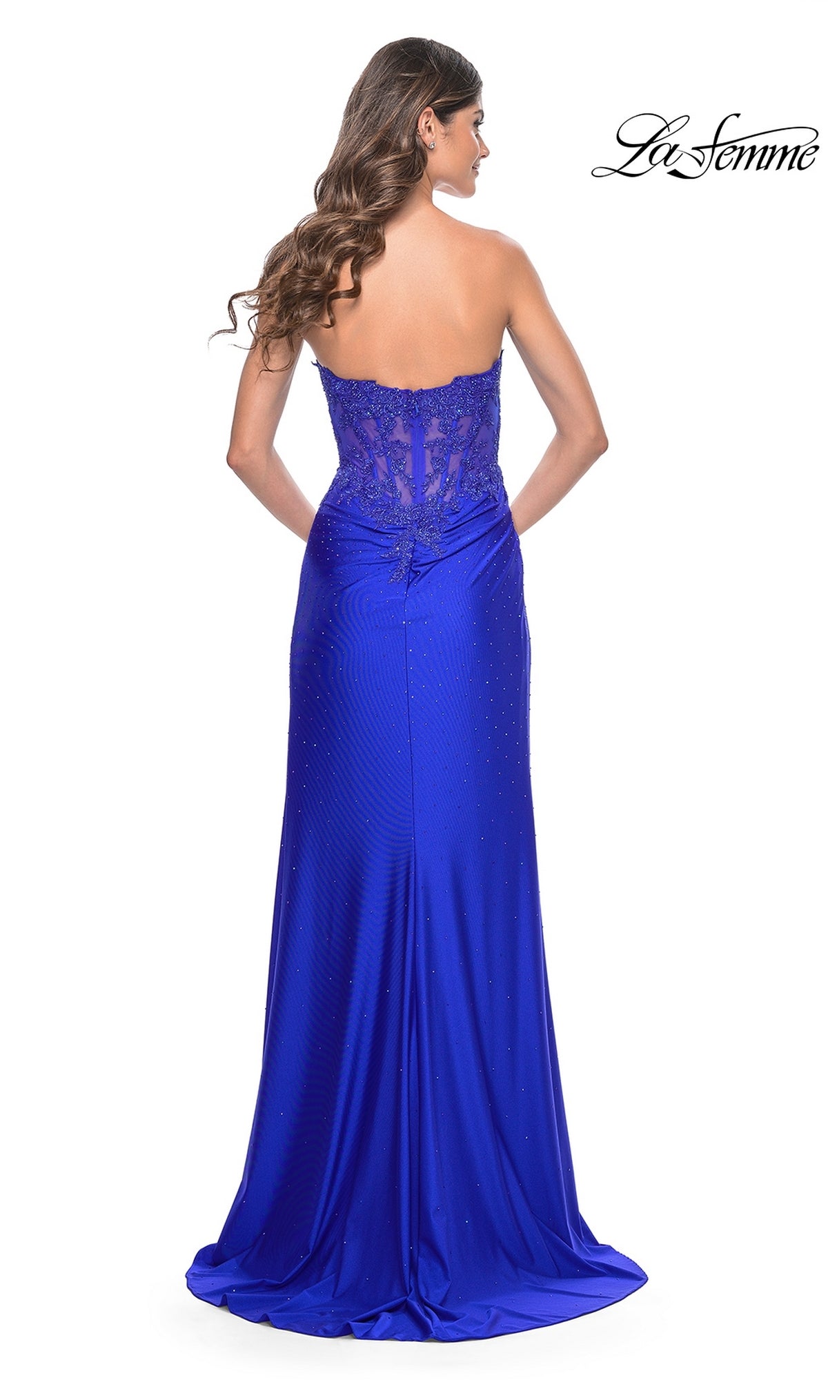 A woman with long wavy hair models the La Femme Sheer-Waist Strapless Long Prom Dress 32011 in royal blue, featuring a sheer lace bodice, subtle embellishments, and elegant lace details. The floor-length gown is shown from the back.