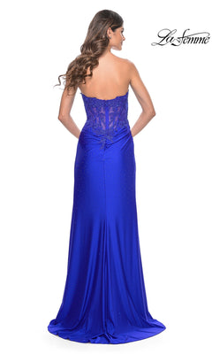 A woman with long wavy hair models the La Femme Sheer-Waist Strapless Long Prom Dress 32011 in royal blue, featuring a sheer lace bodice, subtle embellishments, and elegant lace details. The floor-length gown is shown from the back.