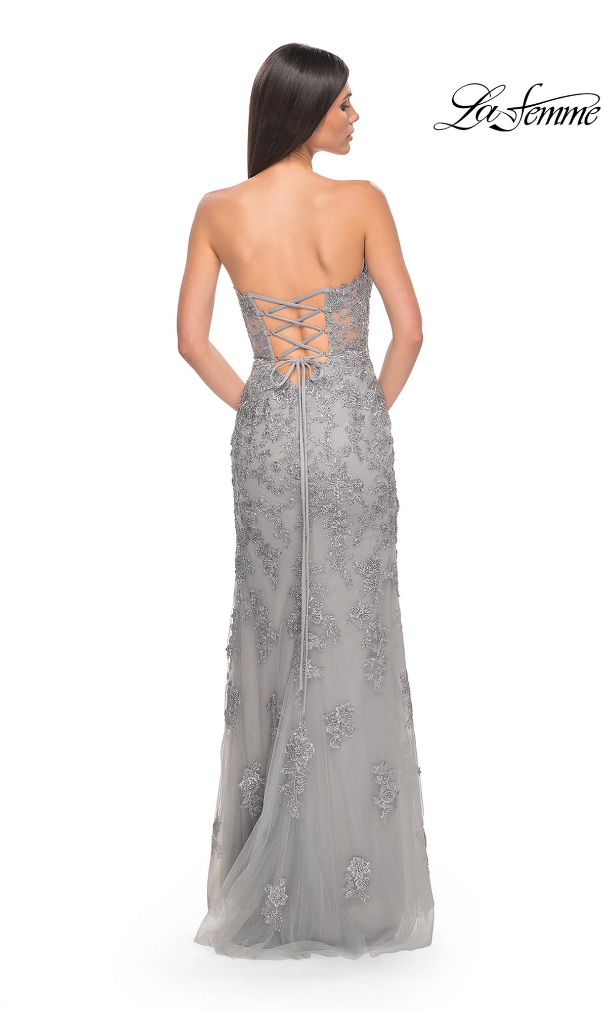 A woman stands with her back to the camera in the La Femme Sheer-Corset Long Prom Dress 32013, a strapless, floor-length gray gown with a lace-up back and floral details. The 