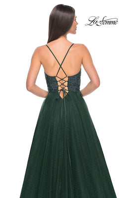 A woman stands with her back to the camera in the La Femme Beaded Long A-Line Prom Dress 32020, featuring a dark green shimmer, thin crisscross straps, a lace-up back, and a sparkling tulle skirt.