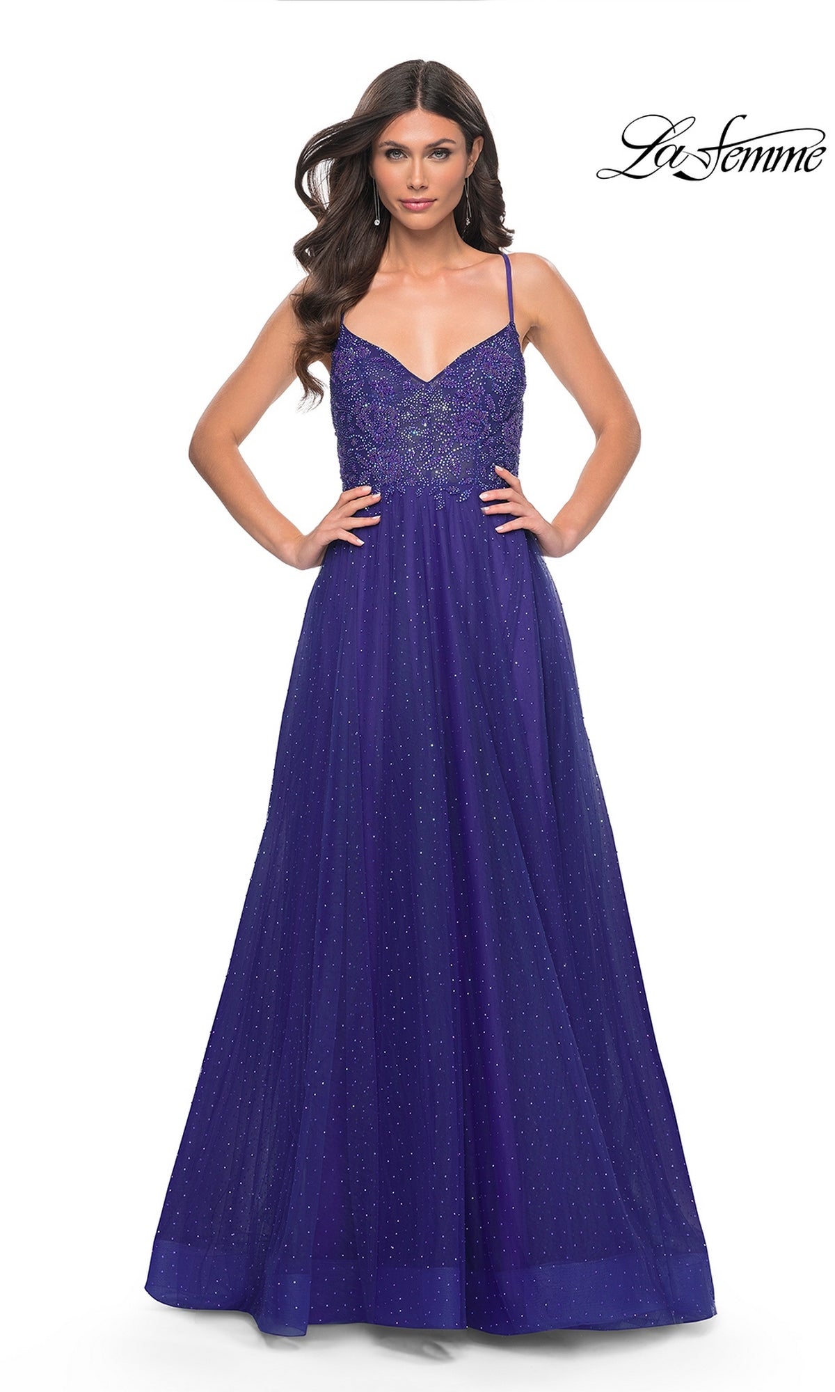 A woman wearing the La Femme Beaded Long A-Line Prom Dress 32020, a sleeveless, floor-length royal blue gown with thin straps and a sparkling bodice, stands with hands on hips. The 