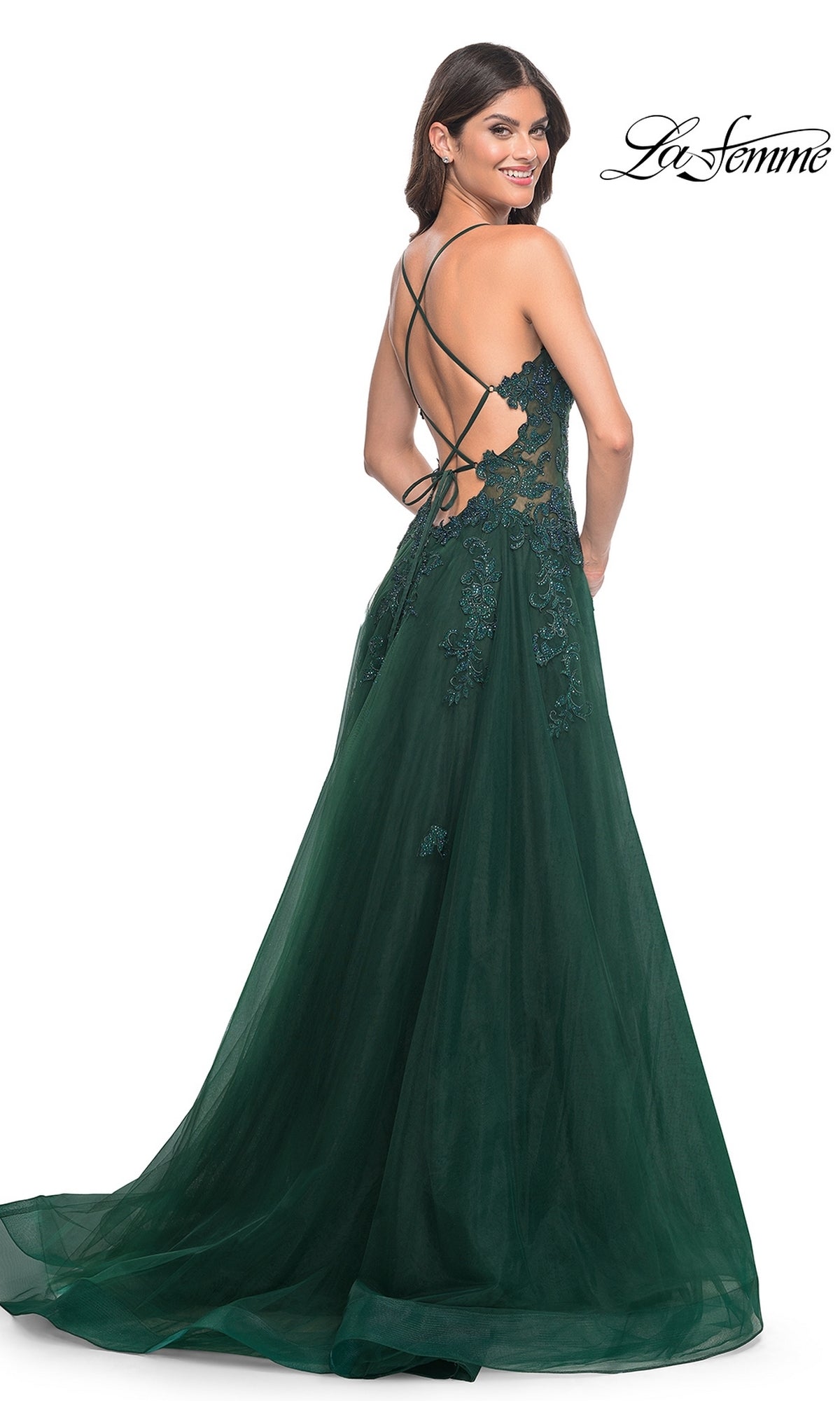 A woman smiles over her shoulder wearing the La Femme Embroidered-Lace Long Prom Gown 32022, a dark green, floor-length dress featuring embroidered lace details, an open back, and crisscross straps.