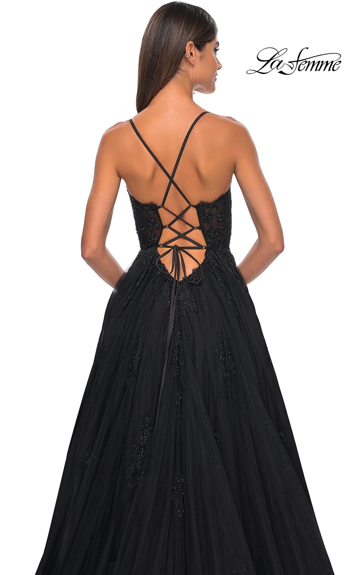A woman stands with her back to the camera wearing the La Femme Sheer-Corset Long A-Line Prom Dress 32028, featuring a black open-back design with lace-up crisscross straps and a flared, subtly embellished skirt for an elegant look.