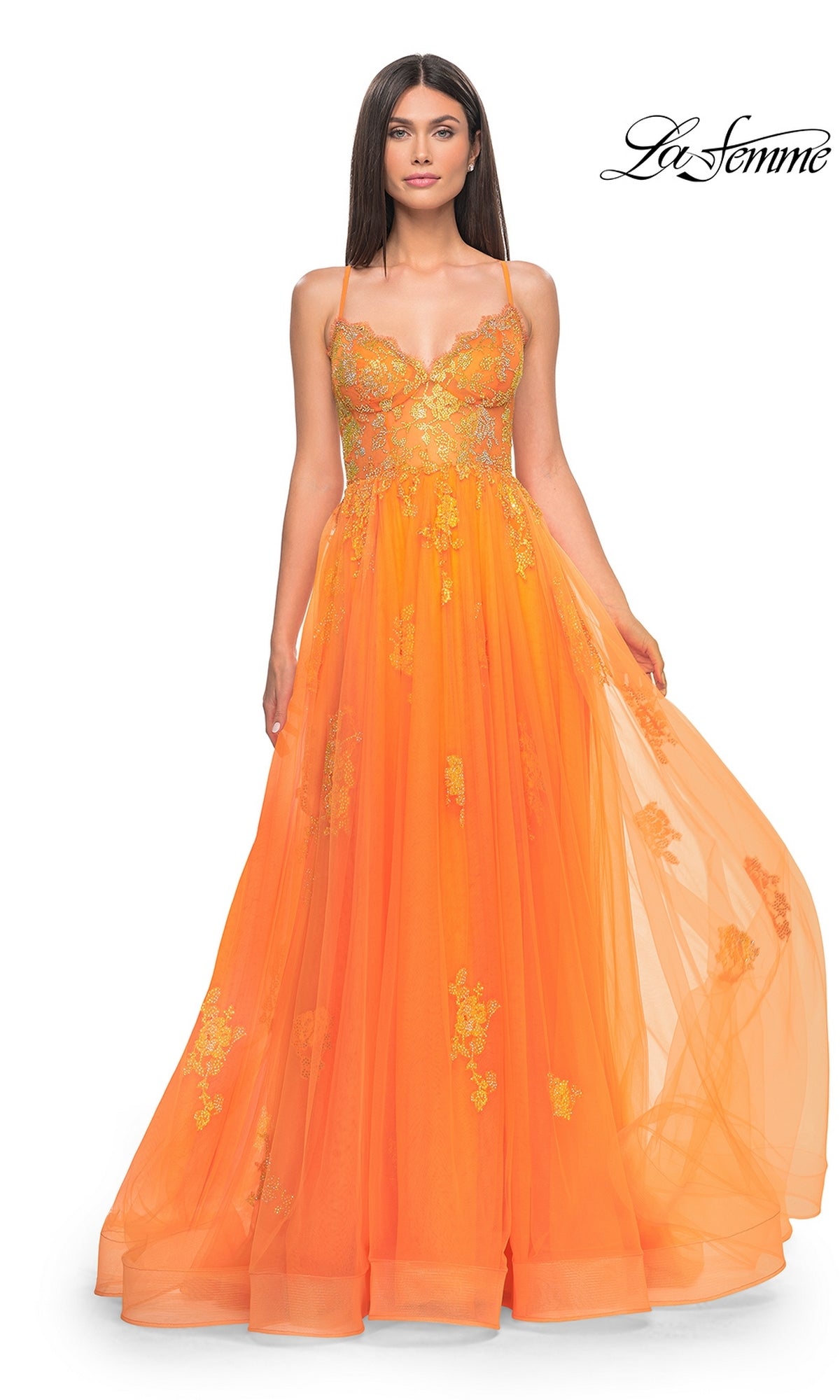 A woman wears the La Femme Sheer-Corset Long A-Line Prom Dress 32028, featuring orange tulle, gold floral embroidery, and thin straps, standing against a white background.