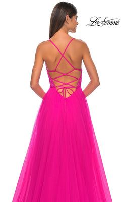 A woman stands facing away, wearing the La Femme Deep V-Neck Long Tulle Prom Gown 32059—a bright pink, floor-length A-line dress with a crisscross strappy open back and long tulle skirt.