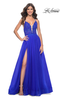 A woman in the La Femme Deep V-Neck Long Tulle Prom Gown 32059, featuring a sleeveless royal blue lace bodice, deep V-neck, high slit, and elegant a-line tulle skirt, smiles against a white background.