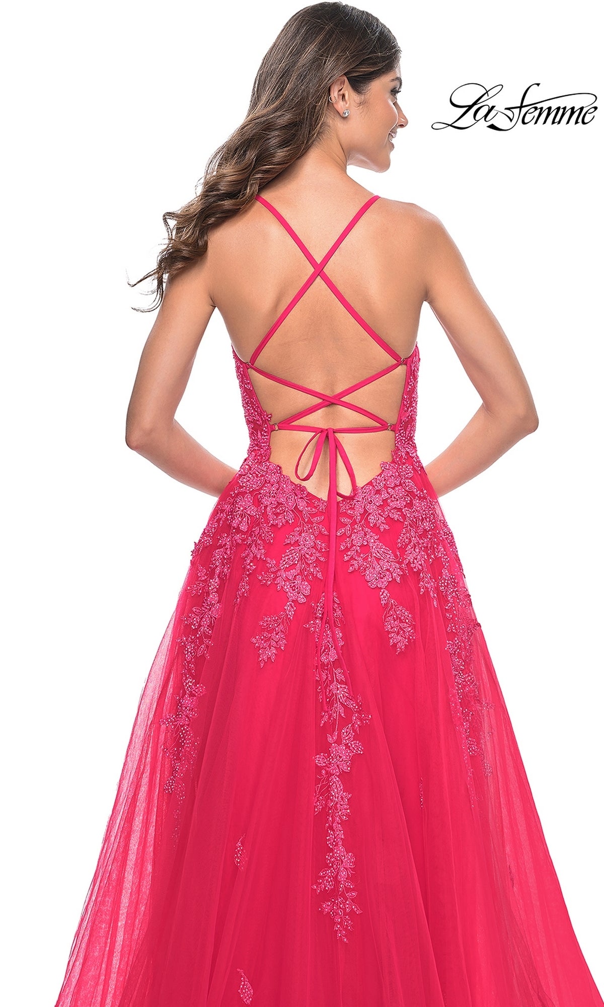 A woman stands facing away in the La Femme Embroidered-Lace Long Prom Dress 32062, featuring a vibrant pink hue, floral lace embroidery, crisscross open-back straps, and a flowing tulle skirt.