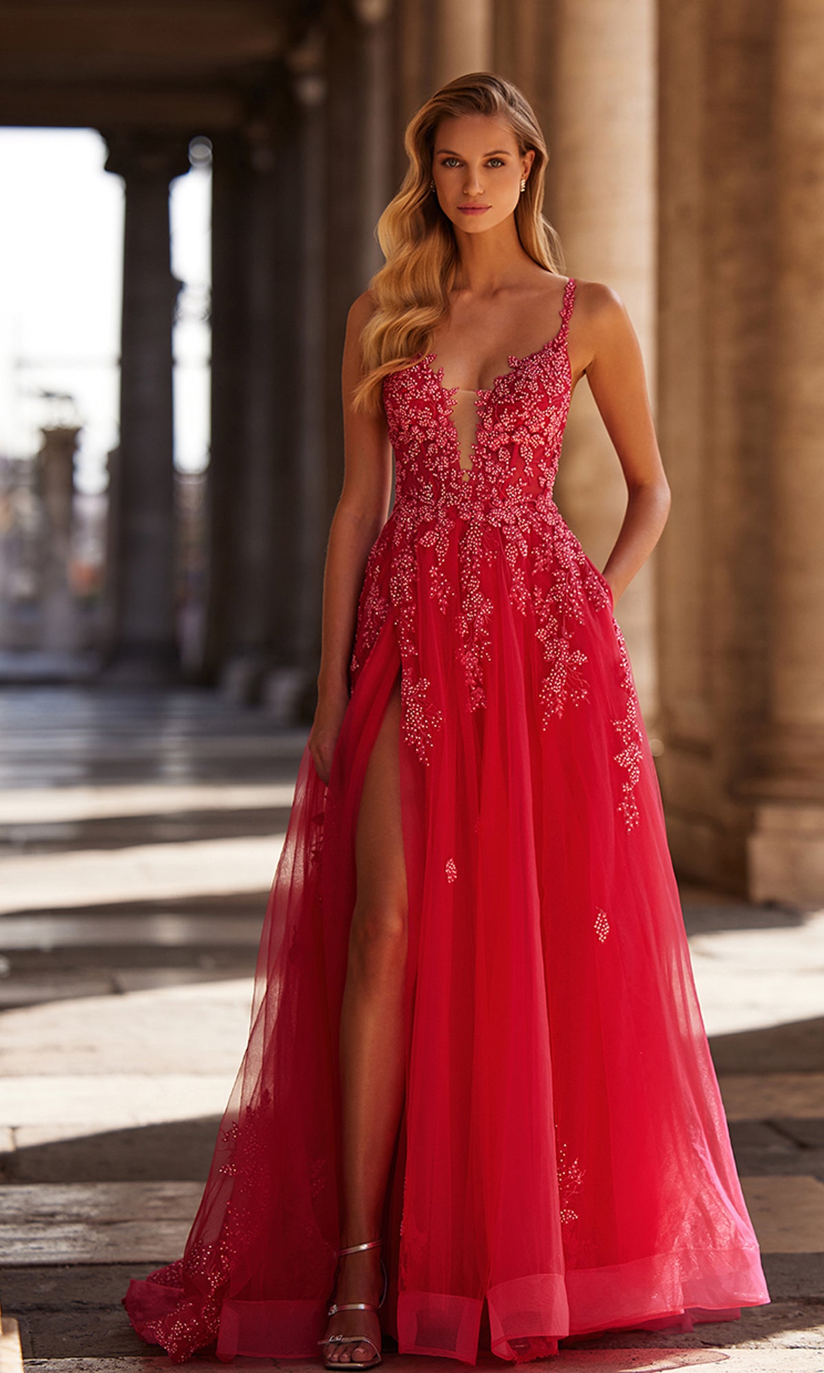 A woman in the La Femme Embroidered-Lace Long Prom Dress 32062, featuring a deep neckline, lace details, and a high slit, stands on a stone walkway with columns behind her.