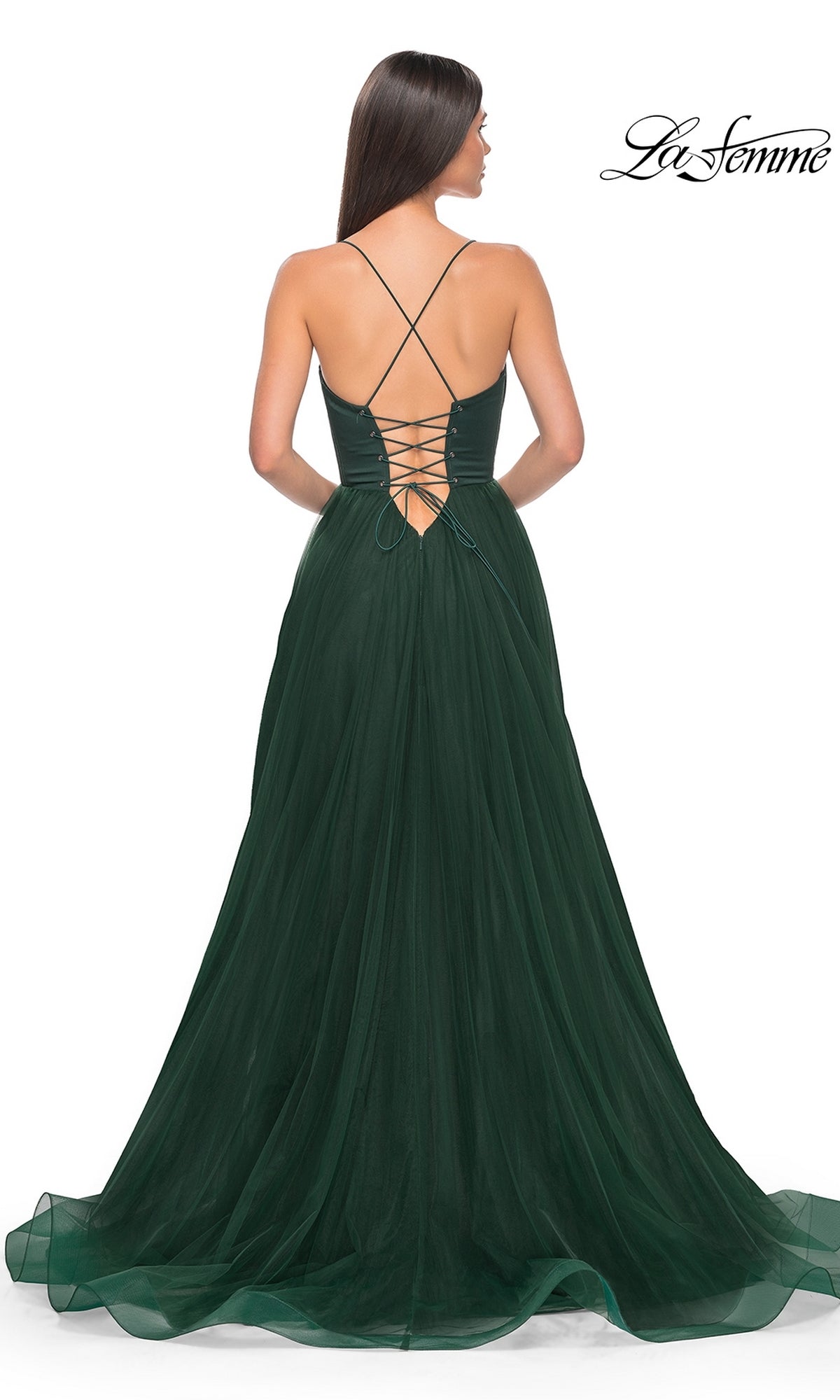 Woman in a floor-length, dark green La Femme Long Sweetheart Prom Ball Gown 32065 with a lace-up back and crisscross straps stands facing away from the camera. La Femme logo visible in the corner.