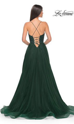 Woman in a floor-length, dark green La Femme Long Sweetheart Prom Ball Gown 32065 with a lace-up back and crisscross straps stands facing away from the camera. La Femme logo visible in the corner.