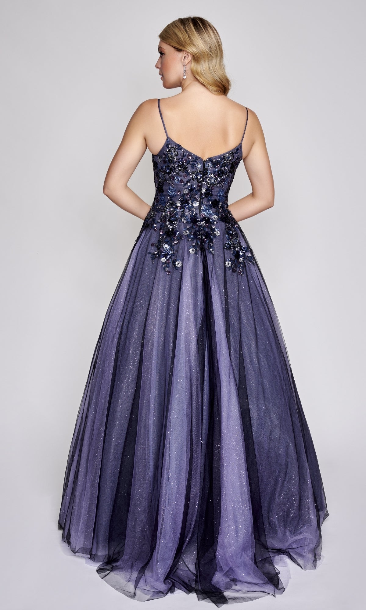 A woman faces away, wearing the Nina Canacci 3206 prom ball gown with thin straps, a floral-embellished bodice, and a floor-length purple and black glitter tulle skirt.