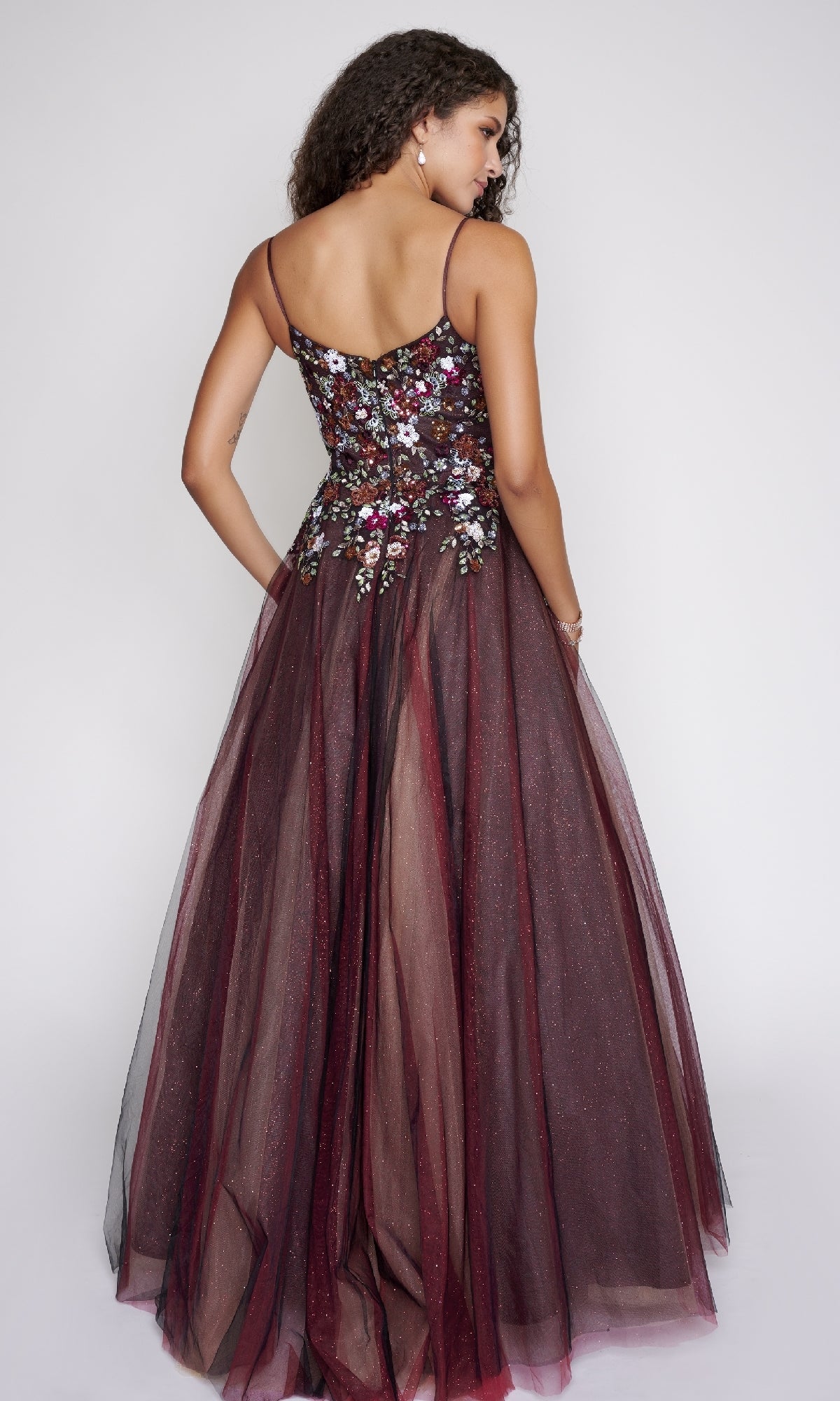 Woman facing away, wearing a sleeveless maroon Nina Canacci 3206 prom ball gown with floral embroidered fitted bodice and a full glitter tulle skirt, set against a plain light background.
