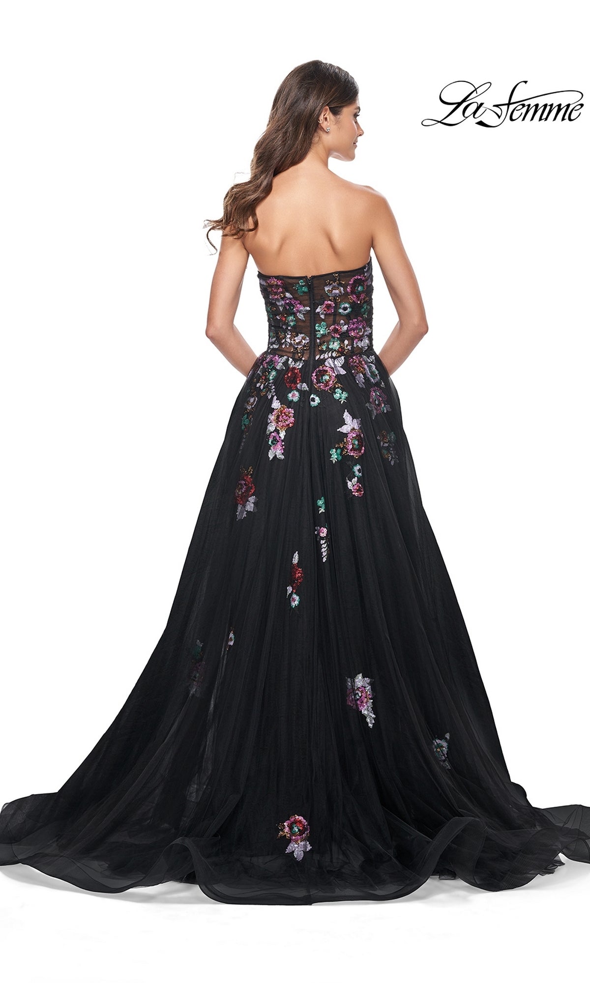 A woman showcases the back and train of the Le Femme Sequin-Floral Long Black Prom Dress 32072, a strapless gown with a sheer overlay and colorful floral embroidery.