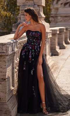A woman stands outdoors by a stone railing, looking to the side, wearing the Le Femme Sequin-Floral Long Black Prom Dress 32072 featuring a strapless neckline, sequin floral details, and a high slit.