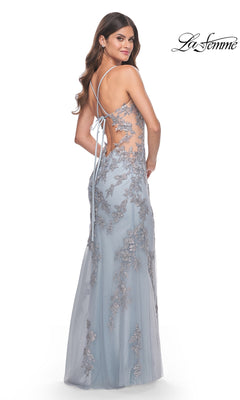 A woman models the La Femme Sheer-Sides Long Blue Prom Dress 32074, featuring silver floral embellishments, embroidered lace, an open lace-up back, and sheer sides as she glances over her shoulder.