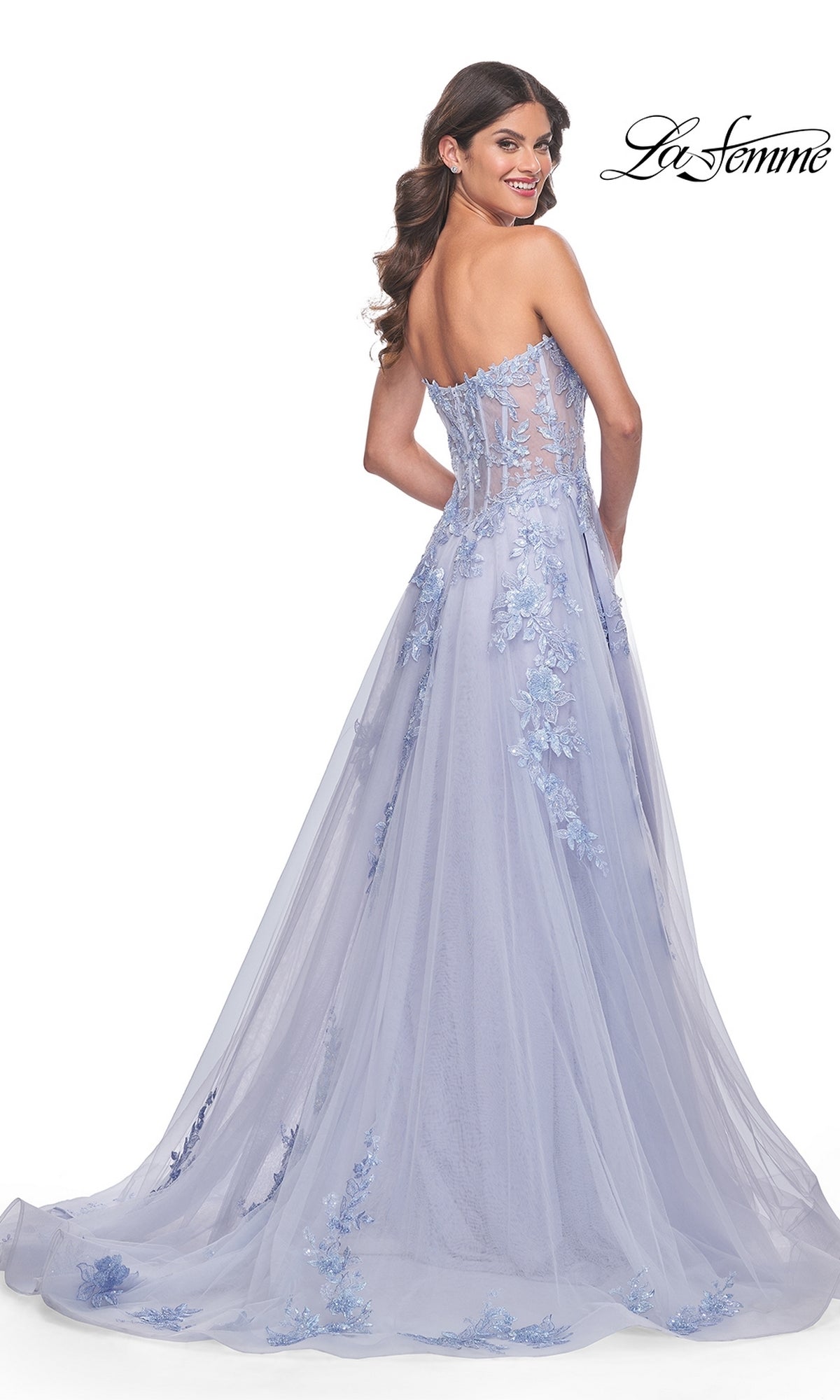 A woman wears the La Femme Strapless Long A-Line Corset Prom Dress 32082 in light purple, featuring floral lace embroidery and a sheer bodice, as she poses and smiles over her shoulder.