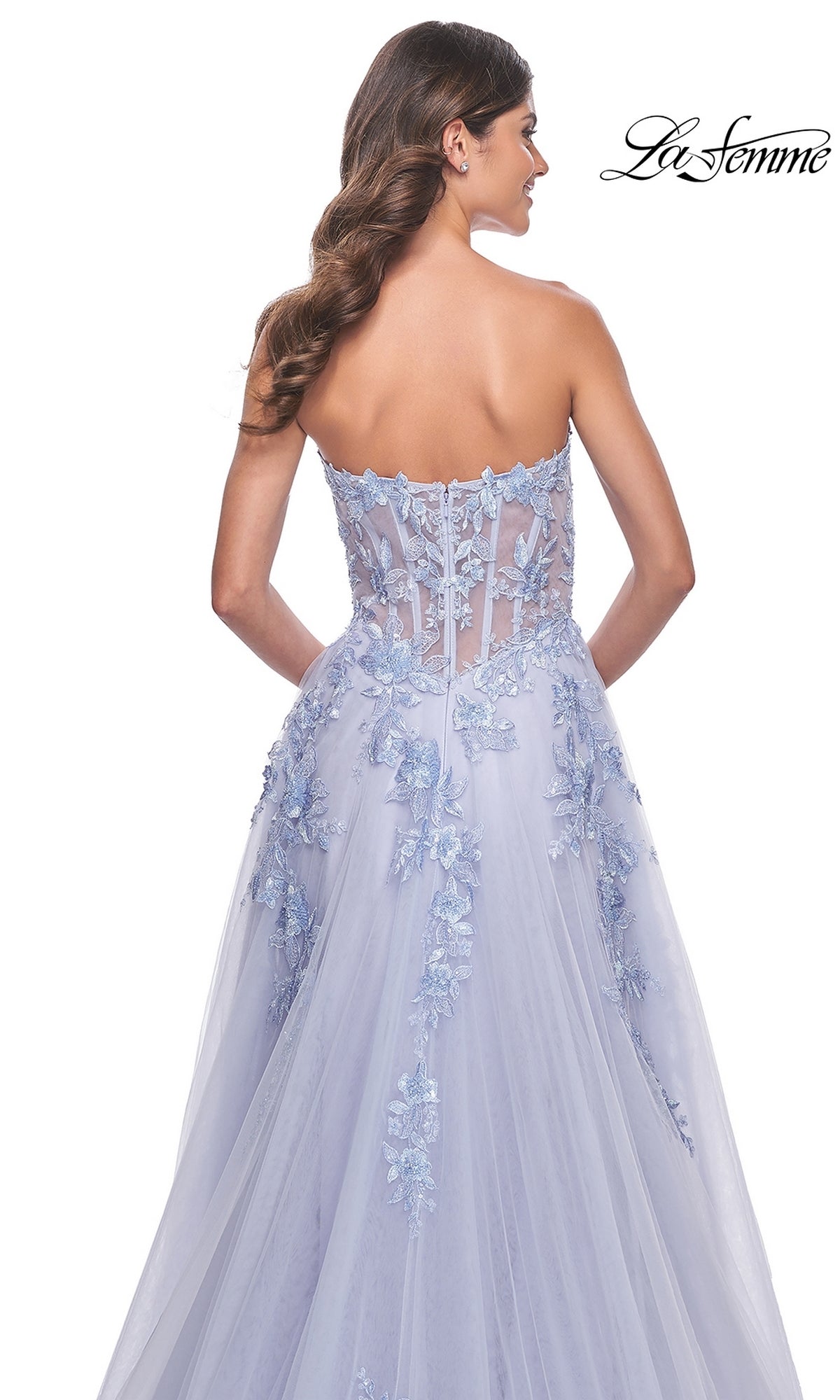 Wearing the La Femme Strapless Long A-Line Corset Prom Dress 32082, a woman stands against a white background, showcasing its sheer corset back and floral lace appliqués for an elegant and graceful look.
