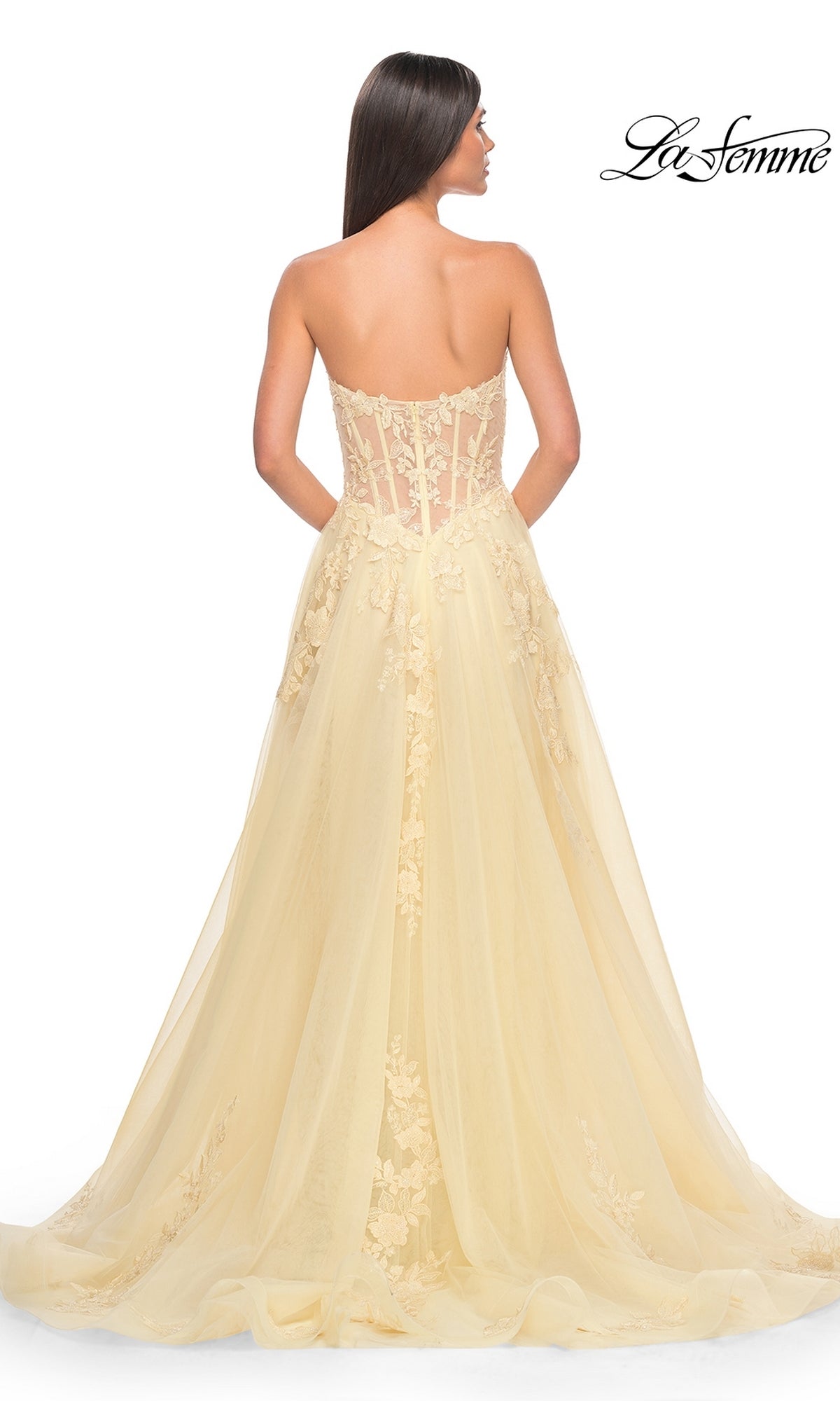 A woman facing away wears the La Femme Strapless Long A-Line Corset Prom Dress 32082 in pale yellow, featuring a sheer floral lace bodice and flowing tulle skirt with train. 