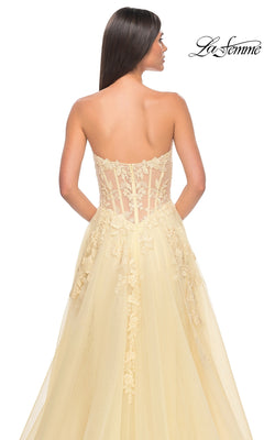 A woman faces away in the La Femme Strapless Long A-Line Corset Prom Dress 32082 with yellow lace embroidery, sheer back, and tulle skirt; "La Femme" logo appears in the top right corner.