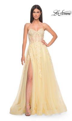 A woman models the La Femme Strapless Long A-Line Corset Prom Dress 32082 in pale yellow with lace embroidery, a sheer bodice, and high slit, standing against a white background. The “La Femme” logo appears in the top right corner.