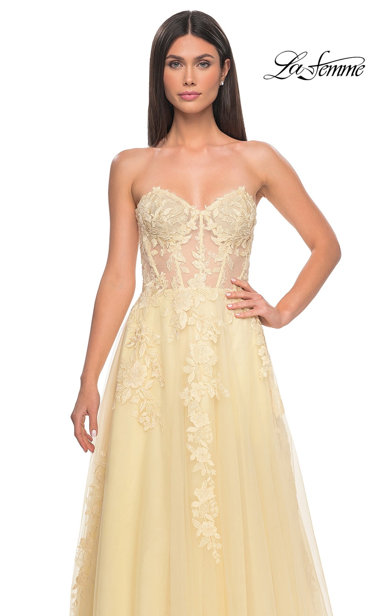 A woman models the La Femme Strapless Long A-Line Corset Prom Dress 32082 in pale yellow, featuring lace embroidery on the bodice and skirt, standing against a white backdrop.