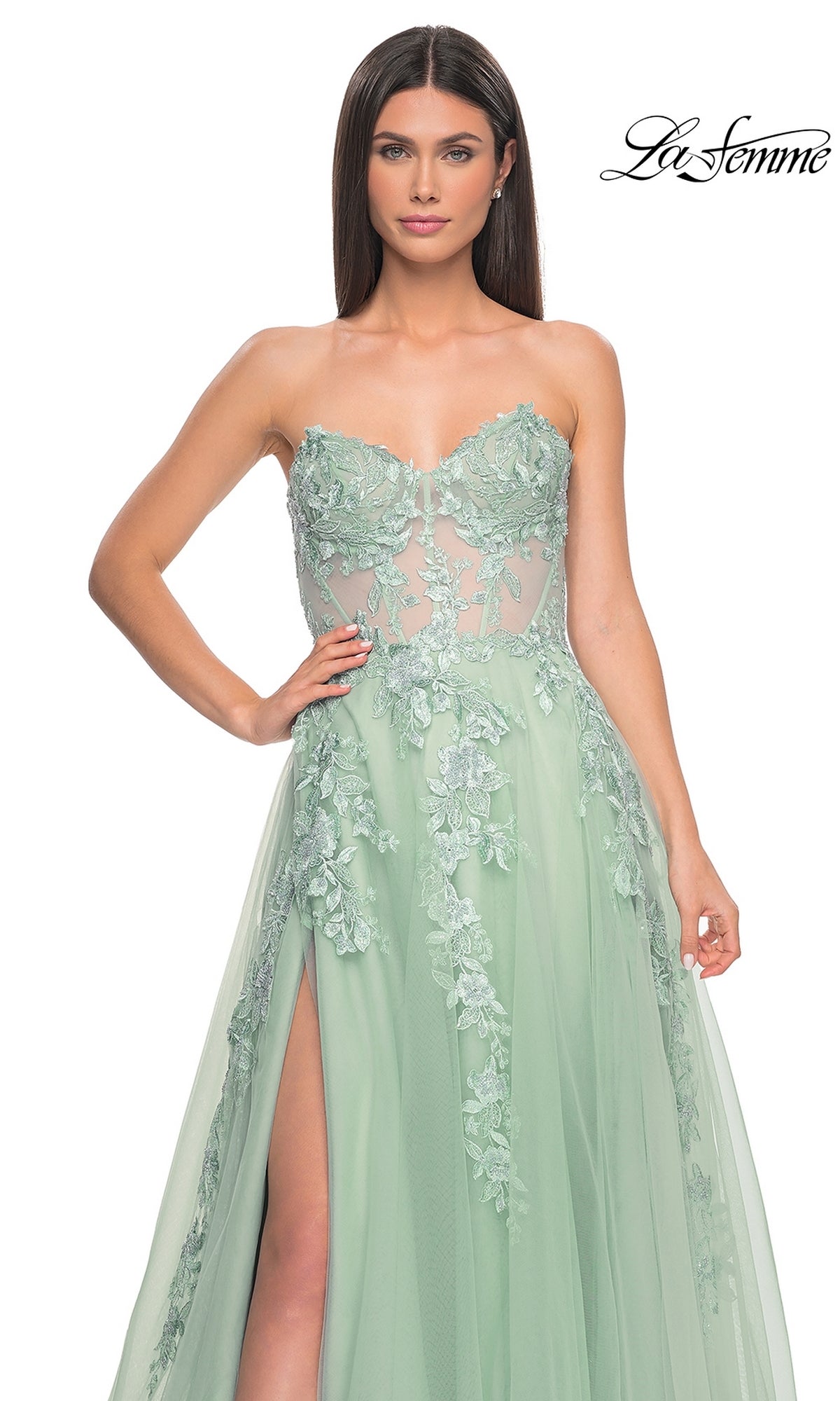 A woman models the La Femme Strapless Long A-Line Corset Prom Dress 32082 in light green, featuring floral lace embroidery, a sheer bodice, and a high slit, standing against a plain white background.