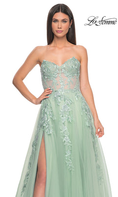 A woman models the La Femme Strapless Long A-Line Corset Prom Dress 32082 in light green, featuring floral lace embroidery, a sheer bodice, and a high slit, standing against a plain white background.