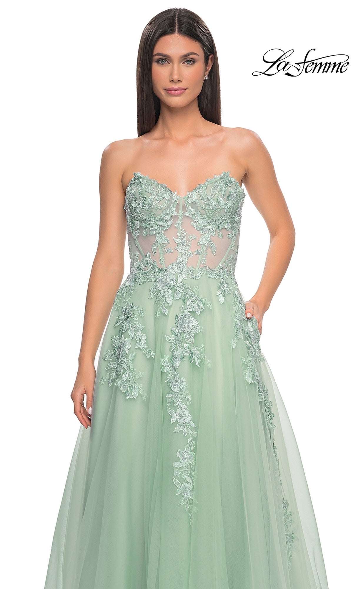A woman wearing the La Femme Strapless Long A-Line Corset Prom Dress 32082, a mint green gown with a sheer bodice and floral lace appliqué, stands against a plain white background.