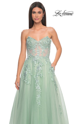 A woman wearing the La Femme Strapless Long A-Line Corset Prom Dress 32082, a mint green gown with a sheer bodice and floral lace appliqué, stands against a plain white background.