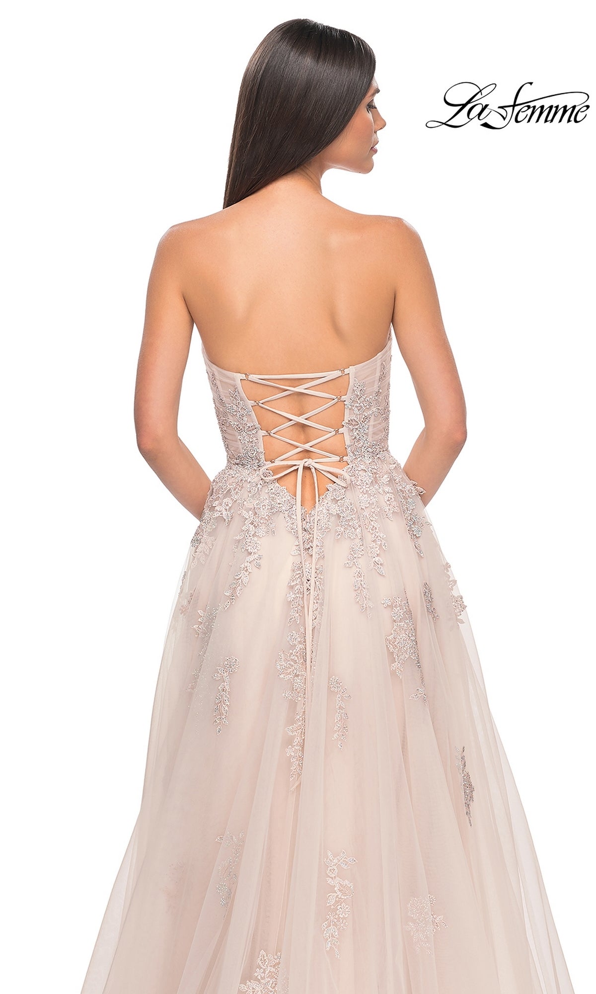 A woman is seen from the back wearing the La Femme Backless Strapless Long Prom Dress 32084, featuring a blush hue, lace-up back, embroidered accents, and a flowing tulle skirt.