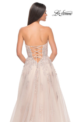 A woman is seen from the back wearing the La Femme Backless Strapless Long Prom Dress 32084, featuring a blush hue, lace-up back, embroidered accents, and a flowing tulle skirt.