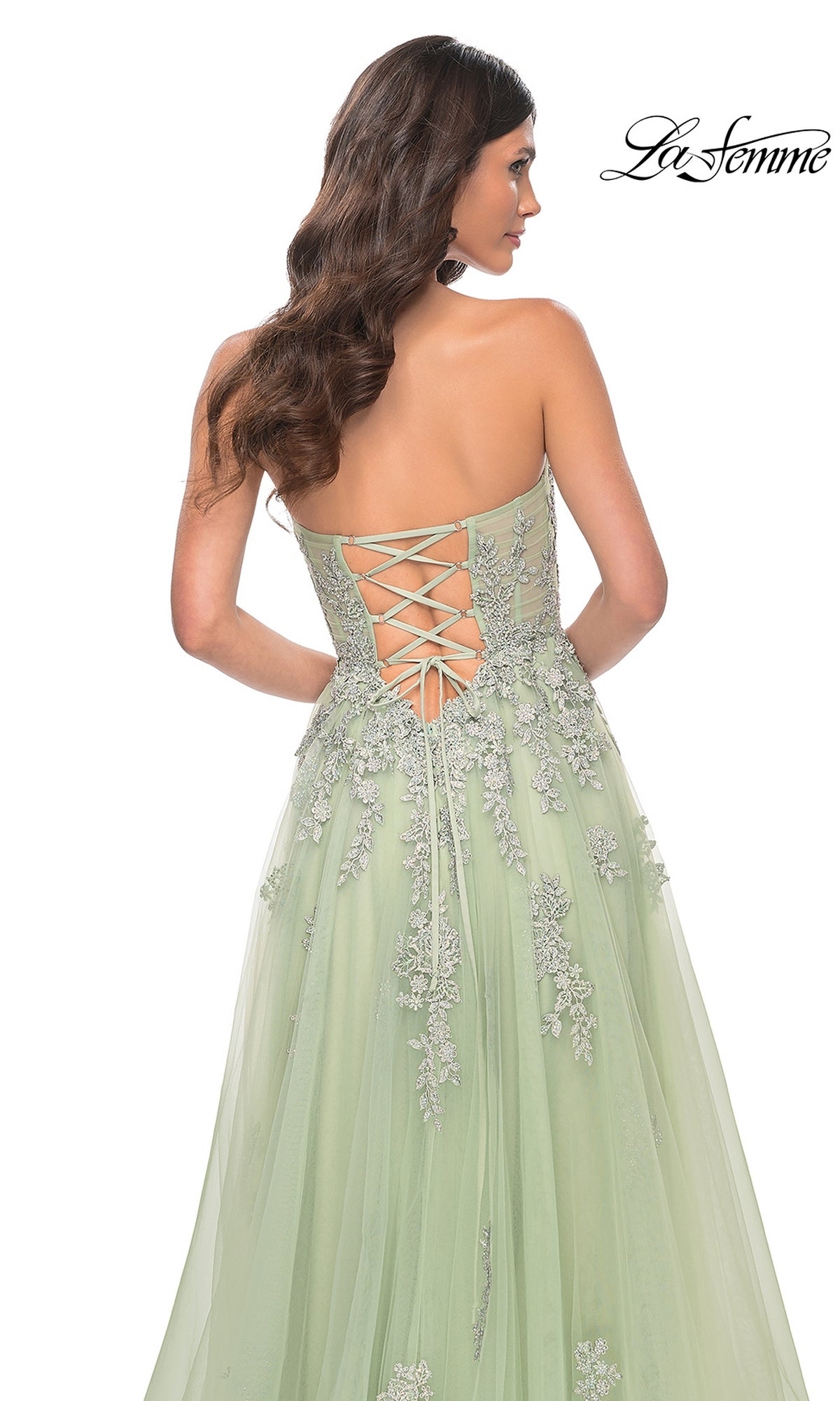 A woman is seen from behind wearing the La Femme Backless Strapless Long Prom Dress 32084 in light green, with a lace-up back, floral embroidered lace accents, and a flowing tulle skirt. The 